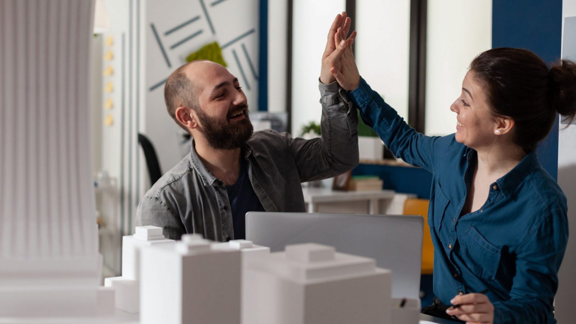 Photo of man and woman high-fiving each other in front of model of building illustrates blog: "What Are the Benefits of Using SBA Financing for Commercial Real Estate?"