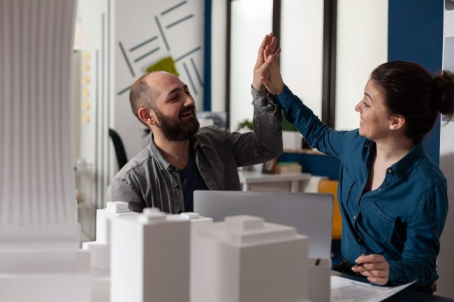 Photo of man and woman high-fiving each other in front of model of building illustrates blog: "What Are the Benefits of Using SBA Financing for Commercial Real Estate?"