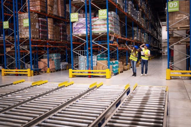 Man and woman wearing safety helmets and vests in a distribution center or warehouse.